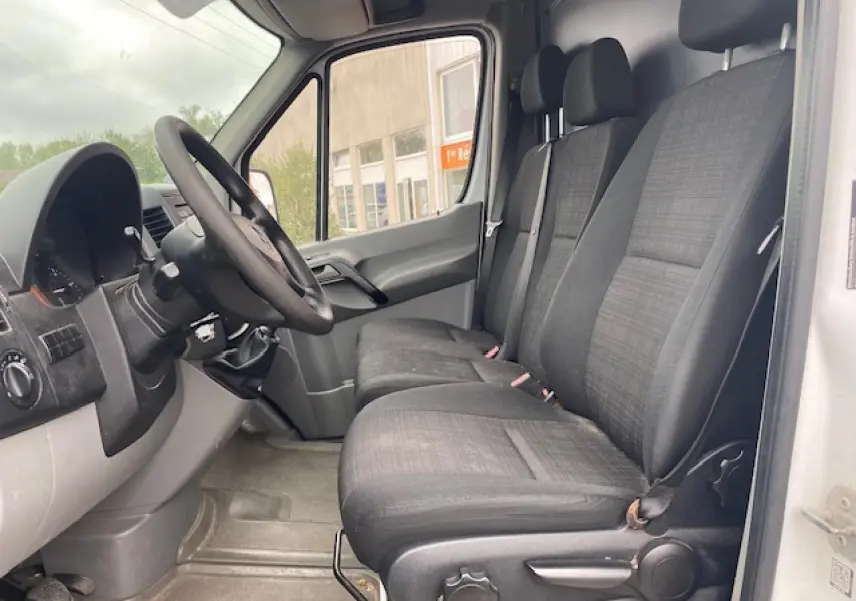 Vue intérieure côté conducteur du Mercedes Sprinter blanc 2015, montrant les sièges avant en tissu gris et le tableau de bord.