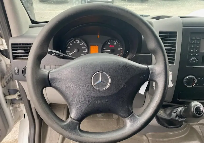 Vue intérieure centrée sur le volant noir Mercedes du Sprinter Fourgon blanc, avec tableau de bord et levier de vitesses manuelle visibles.