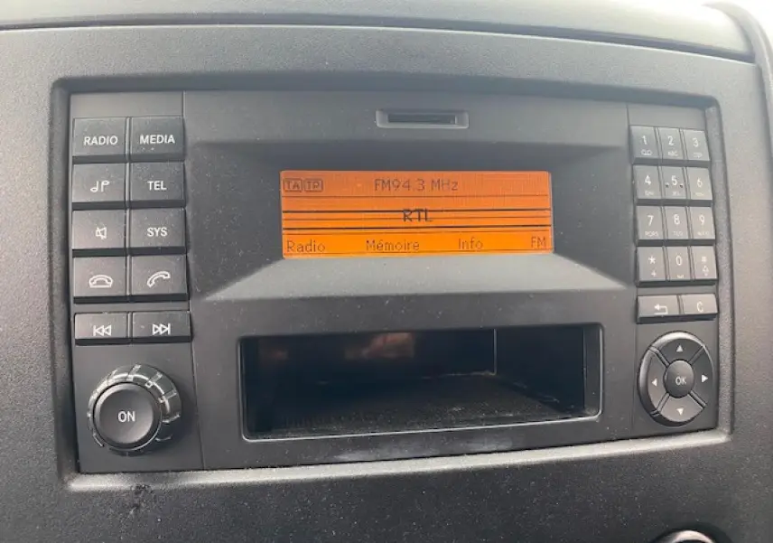 Gros plan sur l’autoradio noir avec écran orange affichant la fréquence FM 94.3 dans un Mercedes Sprinter blanc de 2015.