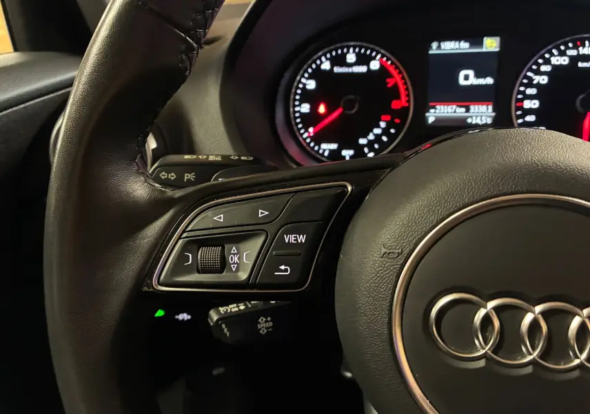 Gros plan sur le volant noir de l'Audi Q2 35 TFSI S-Line avec commandes intégrées et tableau de bord éclairé en arrière-plan.