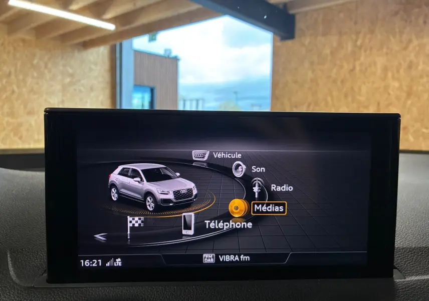 Écran multimédia montrant un Audi Q2 gris en vue 3/4 avant avec menu de sélection des médias et téléphone.