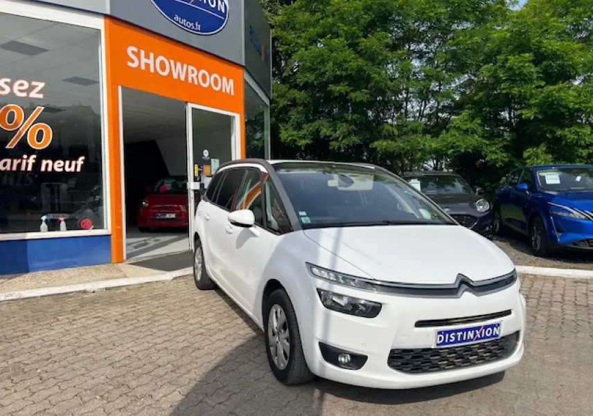 Citroën Grand C4 Picasso blanc en 3/4 avant droit devant un showroom avec végétation en arrière-plan.