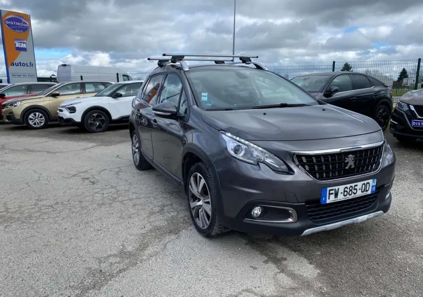 Peugeot 2008 gris platinium vue 3/4 avant droit avec barres de toit aluminium sur parking.