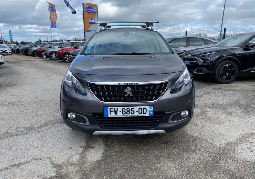 Peugeot 2008 gris platinium vue de face avec barres de toit aluminium sur un parking extérieur.
