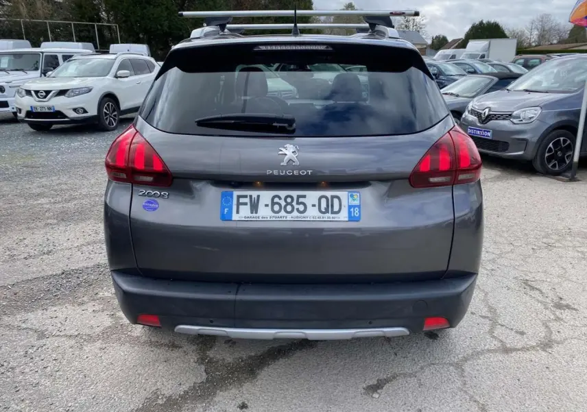 Vue arrière d'un Peugeot 2008 gris platinium avec barres de toit aluminium et caméra de recul visible sur un parking.