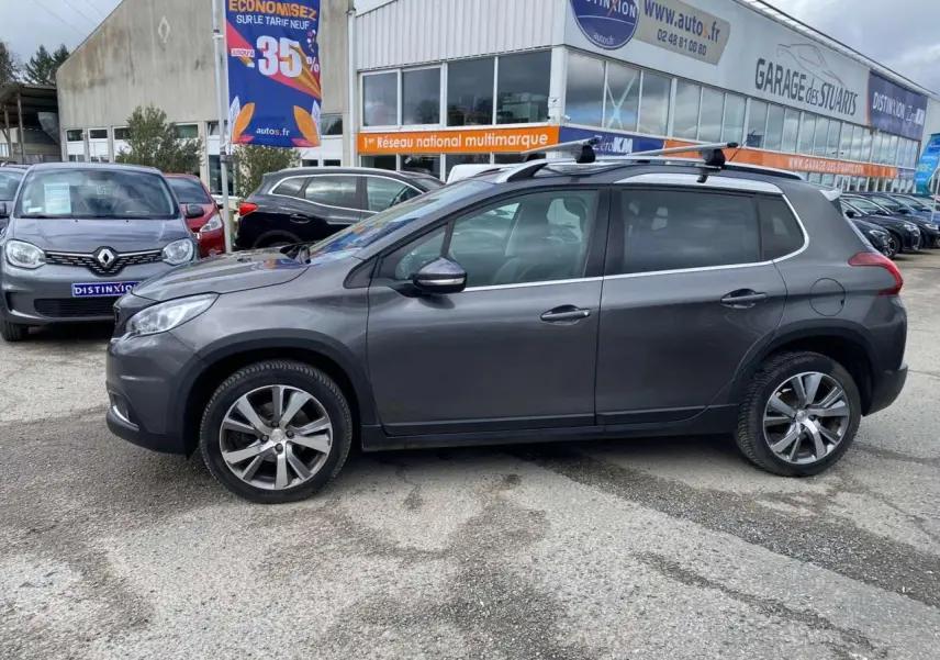 Peugeot 2008 gris platinium vue de profil côté gauche avec barres de toit aluminium sur parking extérieur.