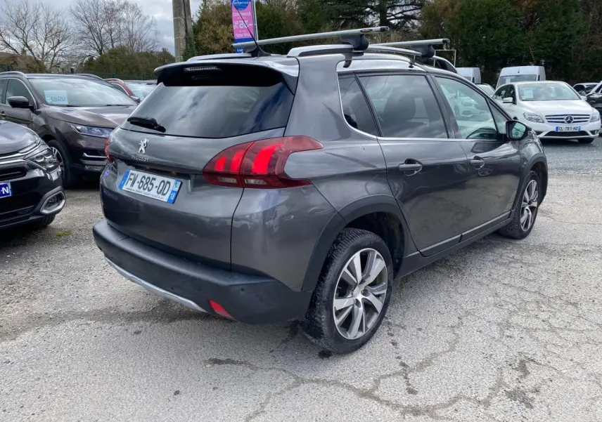 Peugeot 2008 gris platinium vue 3/4 arrière droit avec barres de toit aluminium sur parking extérieur.
