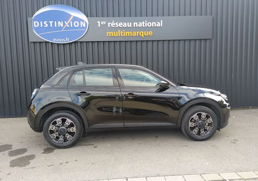 Vue de profil côté gauche d'une FIAT 600 noire 2025 avec jantes noires à motifs blancs, garée devant un mur gris.