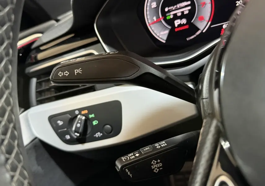 Gros plan sur la commande de clignotants et régulateur de vitesse, tableau de bord numérique partiellement visible, intérieur noir.