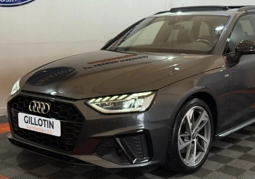 Audi A4 35 TFSI gris foncé vue 3/4 avant droit, avec calandre hexagonale noire et phares LED allumés.