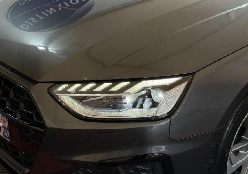 Gros plan sur le phare avant droit et la calandre d'une Audi A4 gris foncé avec jante alliage visible.