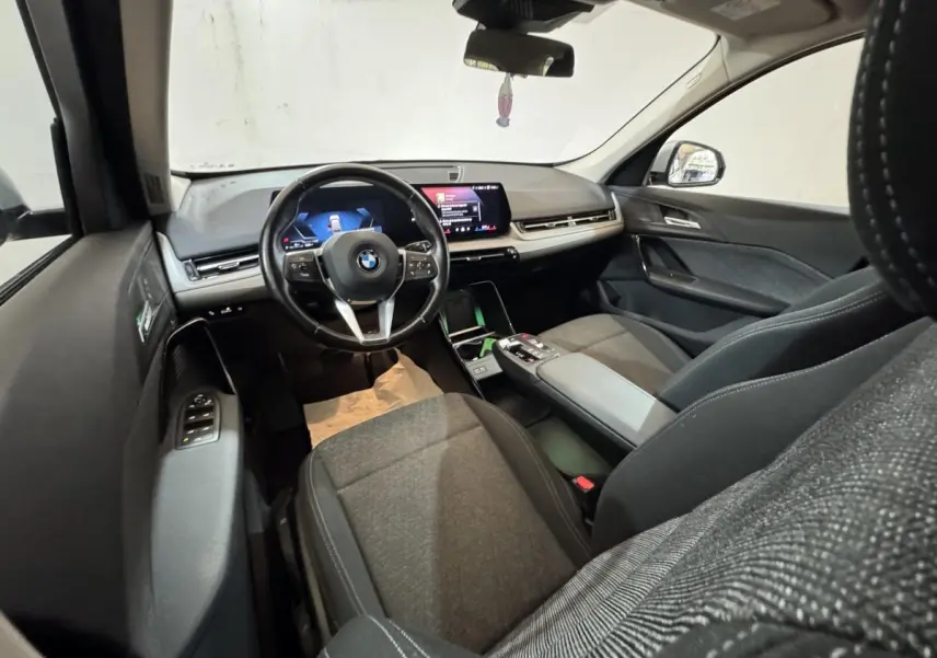 Intérieur BMW X1 2024 vu côté conducteur, sièges gris, volant cuir multifonctions et écran tactile central.