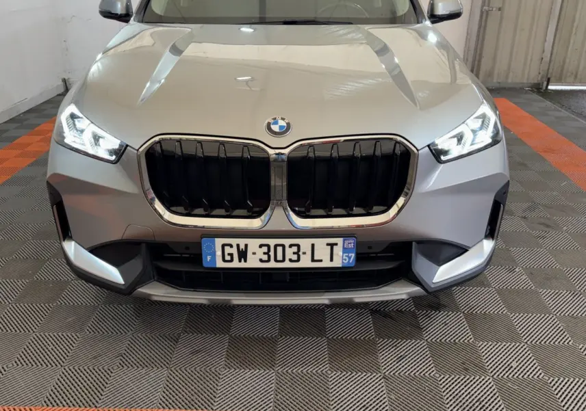 Vue frontale d’un BMW X1 gris foncé 2024 avec calandre noire et phares LED allumés dans un garage.