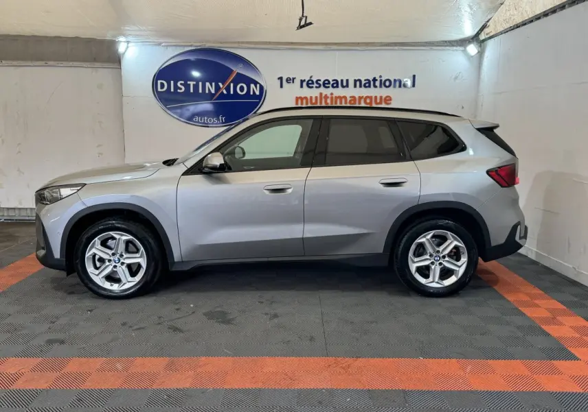 Vue de profil côté gauche d'un BMW X1 gris foncé 2024 dans un showroom avec logo Distinxion en arrière-plan.