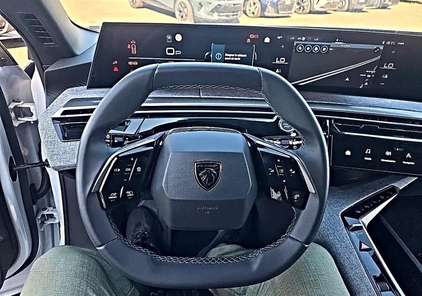 Vue intérieure centrée sur le volant cuir noir et les écrans digitaux du tableau de bord du Peugeot 5008 Hybrid 145 blanc.