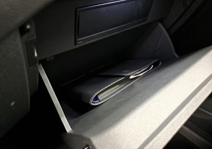 Gros plan de la boîte à gants ouverte du Volkswagen T-Cross 2025, contenant une pochette noire, intérieur sombre.