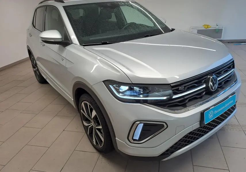 Volkswagen T-Cross 1.0 TSI R-Line 2025 en Reflet d'argent, vue 3/4 avant avec phares allumés et jantes distinctives.