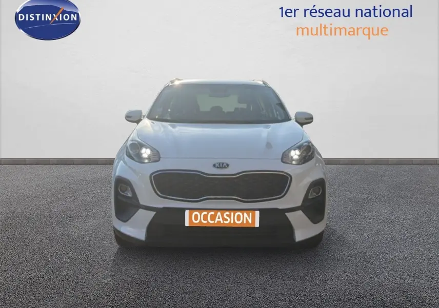 Vue frontale d'un Kia Sportage blanc nacré métal avec phares allumés et plaque "OCCASION" visible.