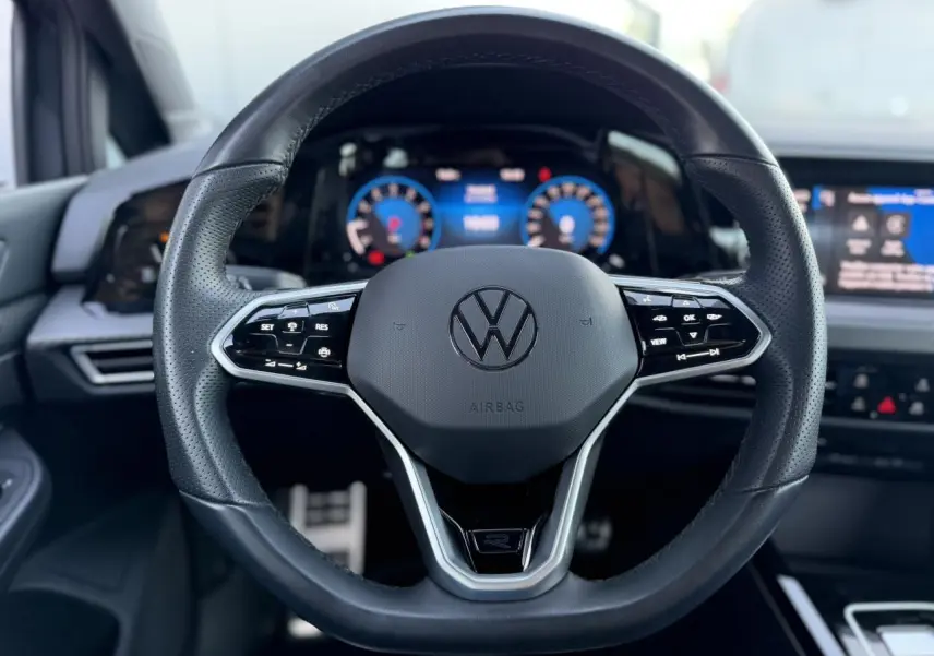 Vue rapprochée du volant cuir multifonction noir avec logo Volkswagen, tableau de bord numérique et écran tactile en arrière-plan.