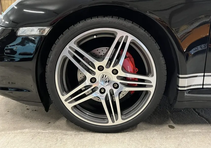 Gros plan sur la jante avant droite de la Porsche 911 Targa noire métallisée avec étrier de frein rouge et bandeau latéral argenté.