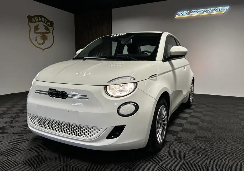 Fiat 500 électrique blanche vue 3/4 avant droit dans un showroom avec phares allumés et jantes acier.