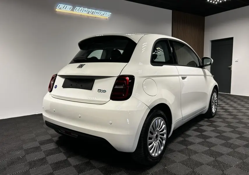 Vue 3/4 arrière droite d'une Fiat 500 électrique blanche 2024 avec jantes acier et feux arrière foncés en intérieur showroom.