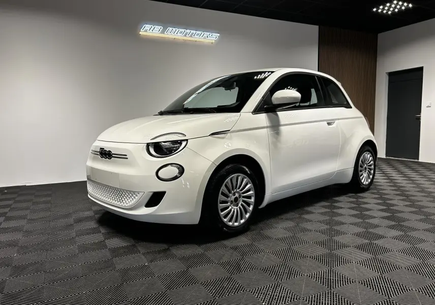 Vue 3/4 avant droite d'une FIAT 500 électrique blanche 2024 avec jantes acier et éclairage LED diurne en showroom.