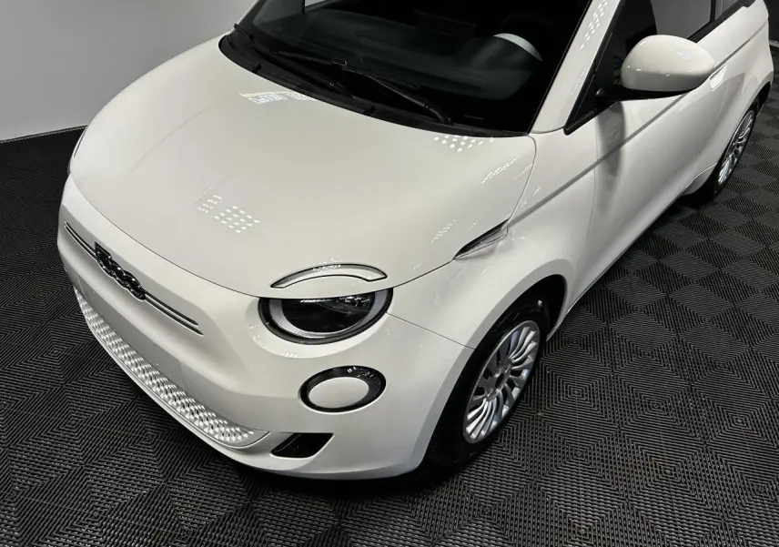 Vue 3/4 avant droit d'une FIAT 500 électrique blanche 2024 avec jantes acier et phares ronds distinctifs.