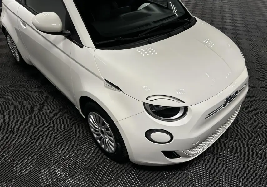 Vue 3/4 avant gauche d'une Fiat 500 électrique blanche 2024 avec jantes acier et phares ronds LED.