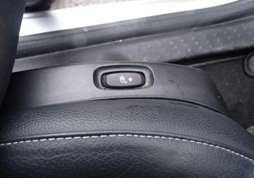 Gros plan sur le bouton de siège chauffant noir avec témoin rouge, entouré de sellerie cuir noir et plastique texturé.