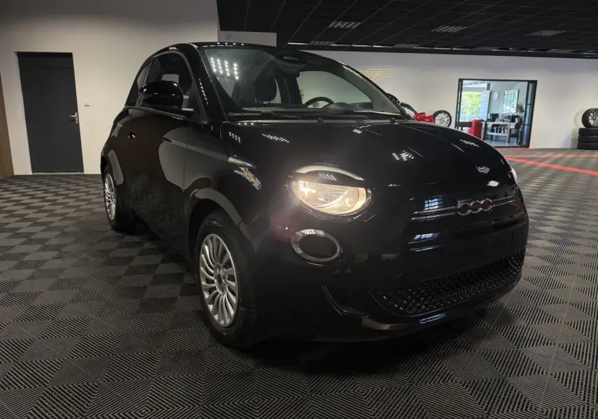 Fiat 500 électrique noire vue en 3/4 avant droit avec phares allumés dans un showroom intérieur.