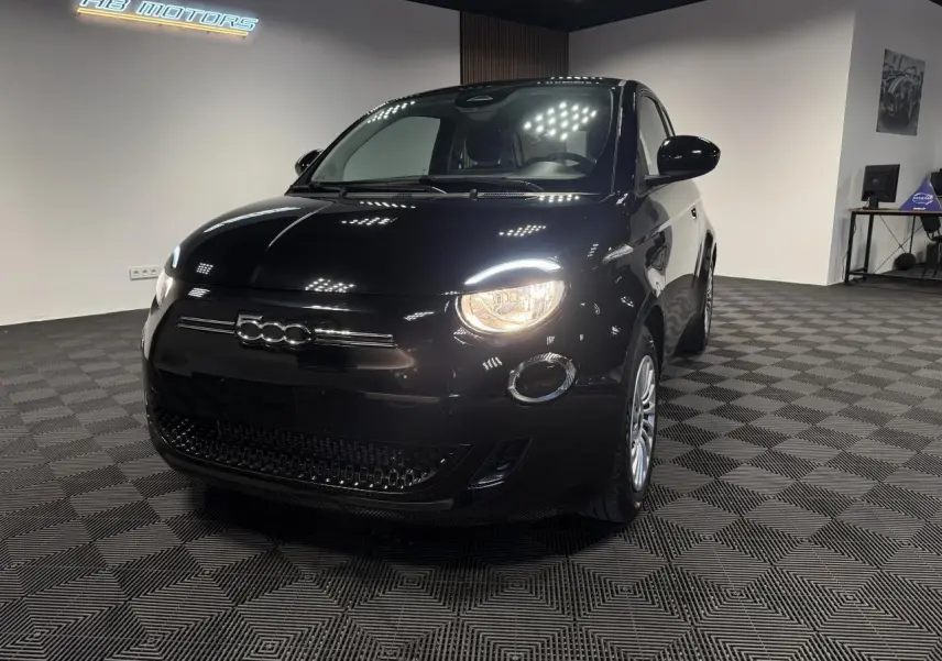 Fiat 500 électrique noire vue 3/4 avant droit avec phares allumés dans un showroom intérieur.