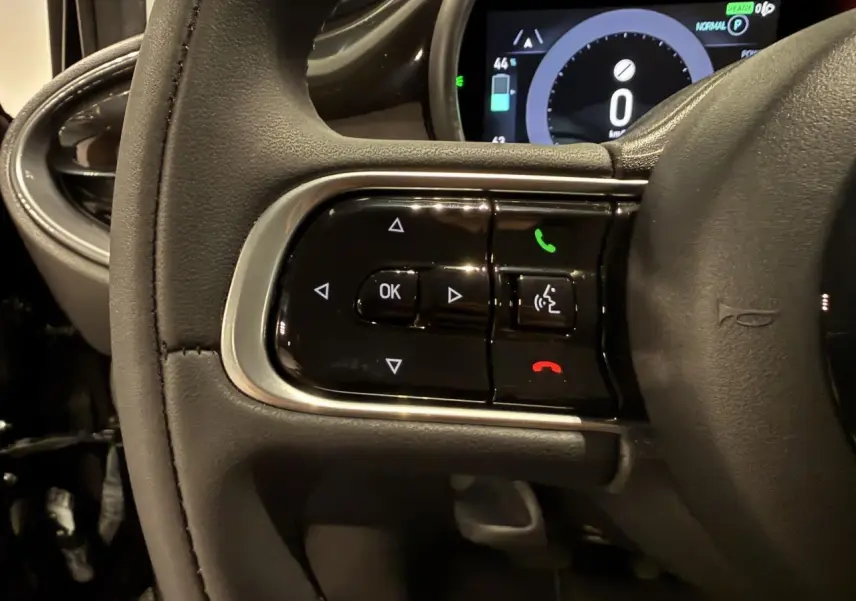Gros plan sur les commandes gauche du volant noir de la Fiat 500 électrique 2024 avec affichage numérique en arrière-plan.