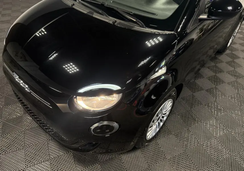 Vue 3/4 avant droit de la Fiat 500 électrique noire 2024 avec phares allumés et jantes acier 15 pouces.