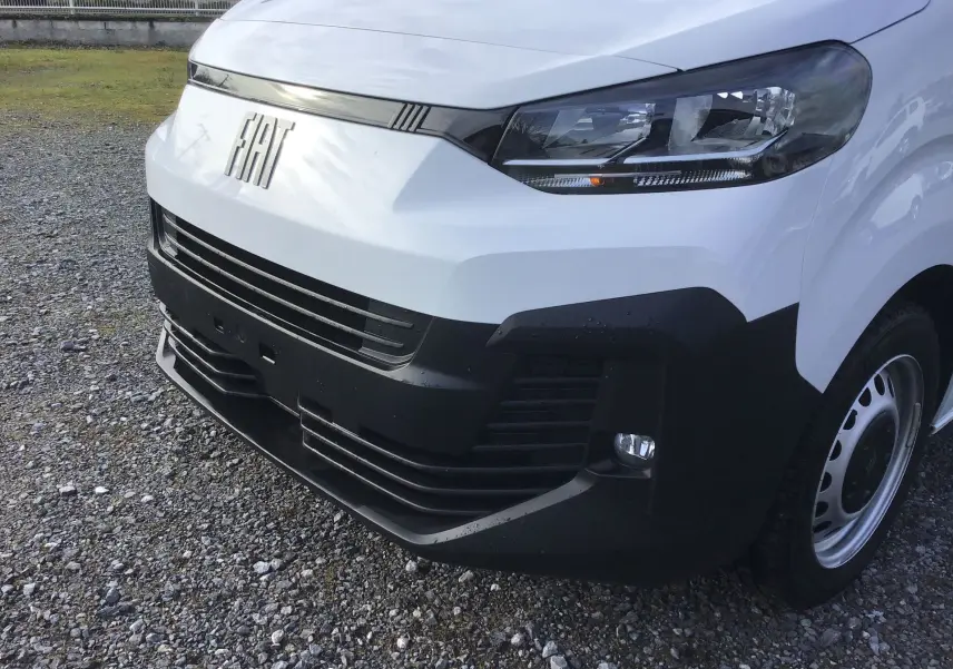 Vue 3/4 avant droit du FIAT Scudo Fourgon blanc avec calandre noire et phares modernes, sur sol gravillonné.