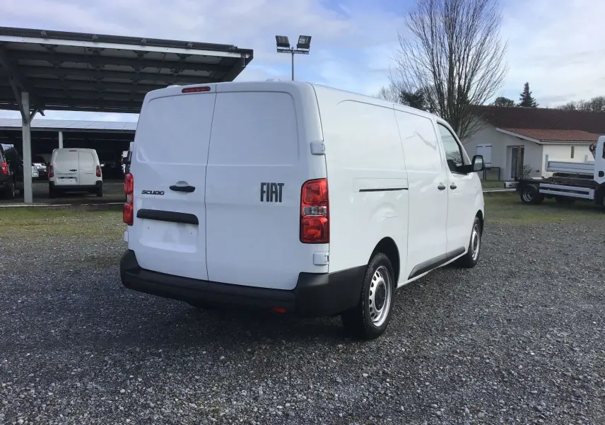 Vue 3/4 arrière droite d’un fourgon FIAT Scudo blanc avec logo FIAT et feux arrière rouges sur un sol gravillonné.