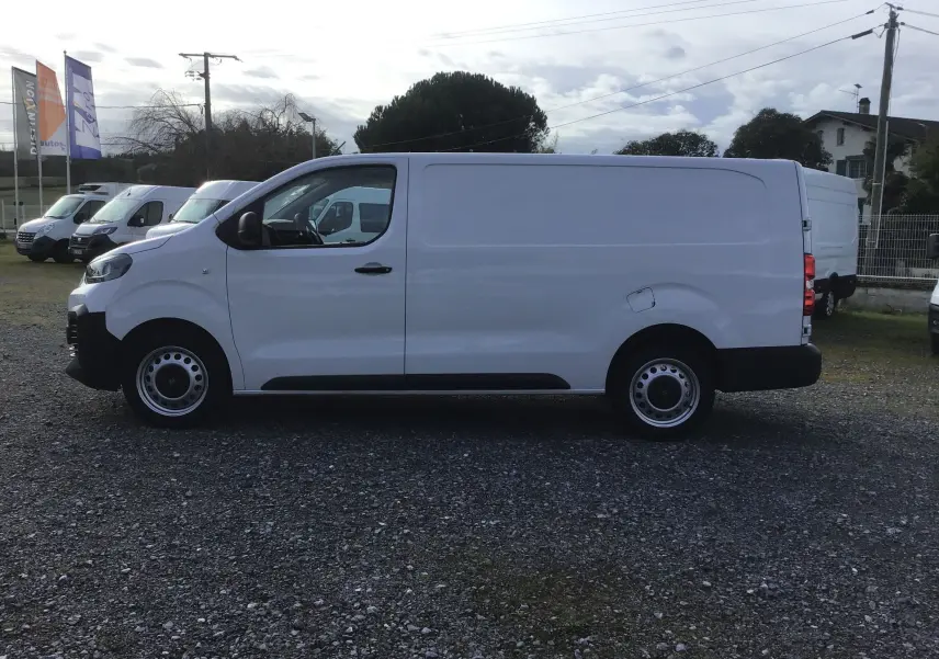 Vue de profil côté gauche d’un FIAT Scudo Fourgon blanc 2024 avec jantes acier et pare-chocs noirs, stationné sur un sol gravillonné.