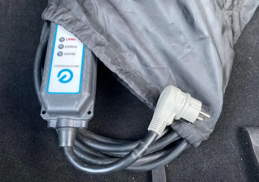 Câble de recharge électrique noir partiellement couvert dans un sac gris, posé sur un tapis de coffre noir.