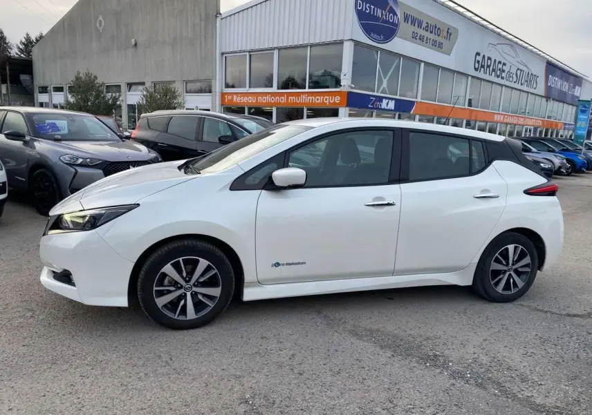 Vue de profil côté gauche d'une Nissan Leaf blanche 40 kWh N-Connecta électrique, stationnée devant un garage multimarque.