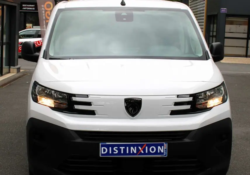 Vue de face du Peugeot Partner Fourgon blanc 2025 avec feux allumés et logo noir distinctif sur la calandre.