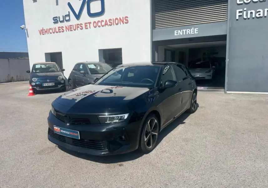 Opel Astra noire 2024 vue 3/4 avant droit, avec jantes alliage 17 pouces et vitres surteintées, stationnée devant un garage.