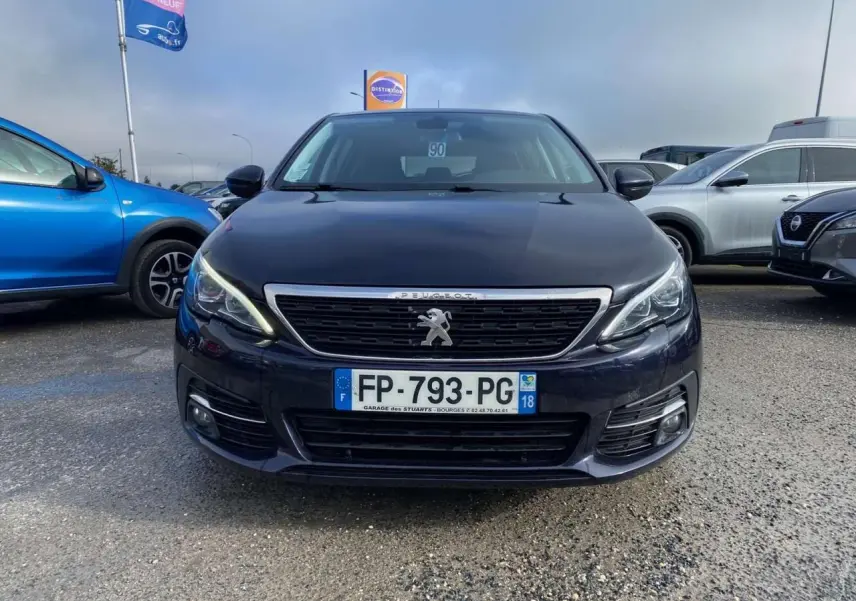 Vue frontale d'une Peugeot 308 bleu foncé avec calandre chromée et feux LED allumés sur un parking extérieur.