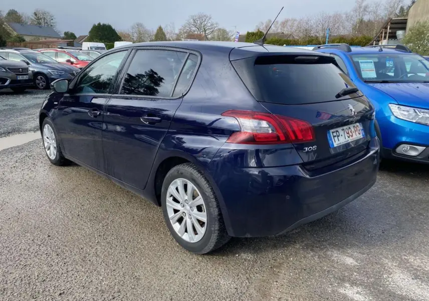 Peugeot 308 bleu foncé vue 3/4 arrière droit, avec feux arrière et jantes visibles sur parking extérieur.