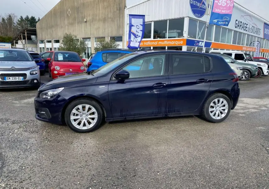 Vue de profil côté gauche d'une Peugeot 308 2018 bleu foncé métallisé, stationnée devant un garage multimarque.