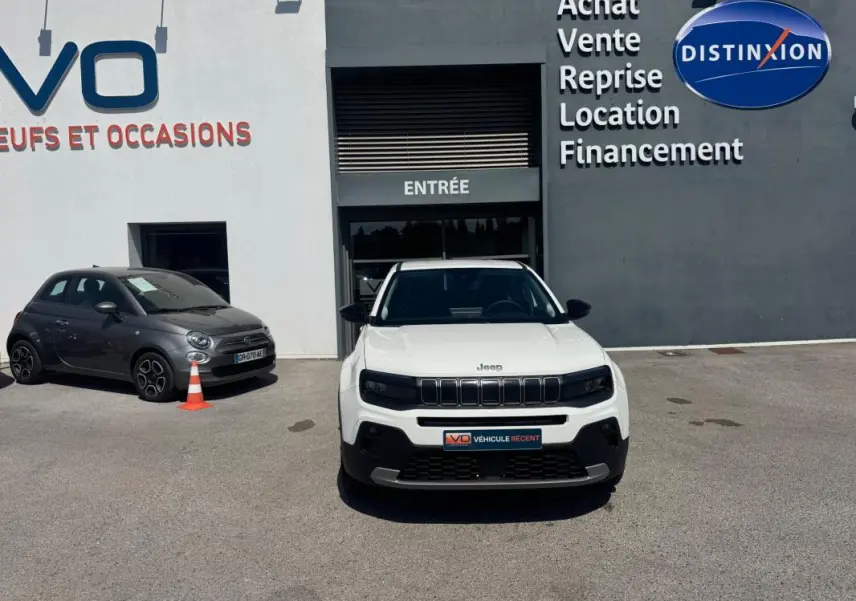 Vue avant d'une Jeep Avenger électrique blanche stationnée devant un bâtiment commercial avec enseignes visibles.