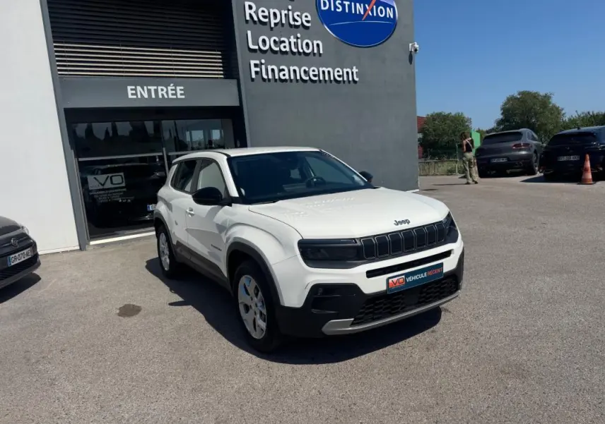 Jeep Avenger électrique blanc en 3/4 avant droit, avec jantes alliage 17 pouces, stationné devant un bâtiment commercial.