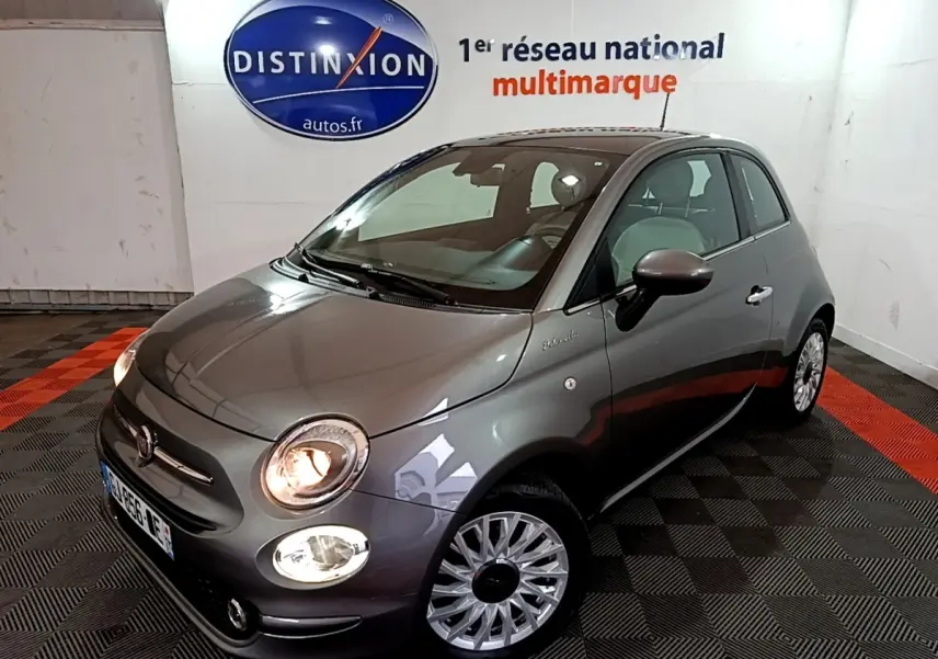 Vue 3/4 avant droite d'une FIAT 500 gris foncé avec phares allumés dans un showroom intérieur.