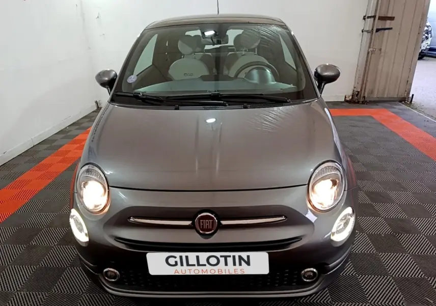 Vue frontale d'une Fiat 500 gris foncé avec phares allumés dans un garage intérieur.