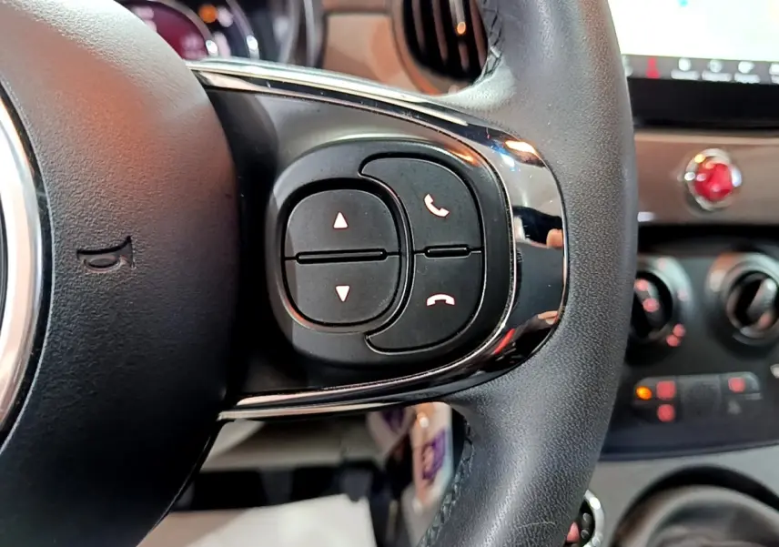 Gros plan sur les commandes du volant cuir noir de la Fiat 500 Dolcevita 2022, avec tableau de bord flou en arrière-plan.