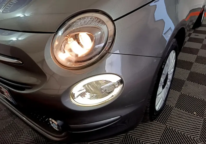 Gros plan sur l'avant droit d'une Fiat 500 gris foncé avec phare principal allumé et jante blanche visible.